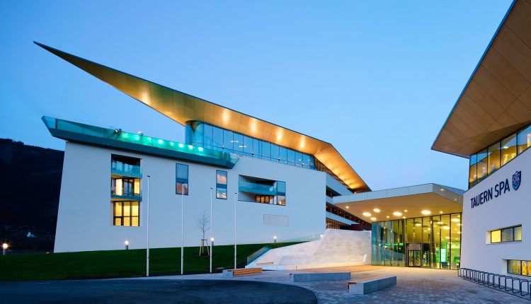 Modernes Gebäude mit kantigen Dächern und großen Fenstern, in der Dämmerung beleuchtet, Eingang mit der Aufschrift TAUERN SPA.