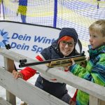Eine Frau und ein Junge zielen mit einem Luftgewehr auf eine verschneite Outdoor-Veranstaltung mit einem Dein Winter Dein Sport-Banner im Hintergrund.
