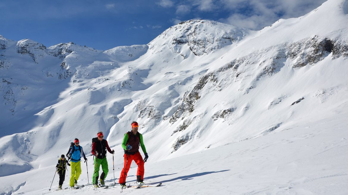 Drei Skifahrer in farbenfroher Ausrüstung erklimmen einen verschneiten Berghang unter einem strahlend blauen Himmel mit schneebedeckten Gipfeln im Hintergrund.