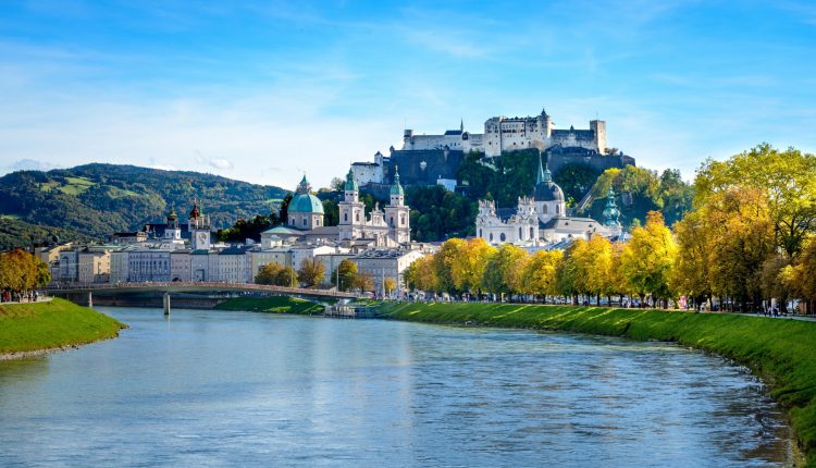 Ein Blick auf das Flussufer von Salzburg, Österreich, mit der Festung Hohensalzburg auf einem Hügel und Herbstbäumen.