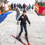 Ein Junge auf Skiern lächelt und zeigt den Daumen nach oben auf einer künstlichen Skipiste, mit Menschen im Hintergrund.