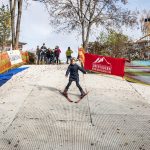 Ein Kind fährt auf Skiern eine trockene Piste hinunter, während mehrere Personen von der Spitze des Hügels aus zusehen.