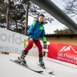 Ein Junge in Skiausrüstung übt auf einer trockenen Skipiste mit Bannern und Bäumen im Hintergrund.