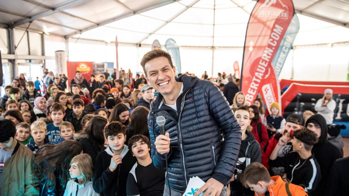 Ein Mann mit einem Mikrofon lächelt bei einer überfüllten Veranstaltung in einer Halle mit vielen Kindern und bunten Bannern.