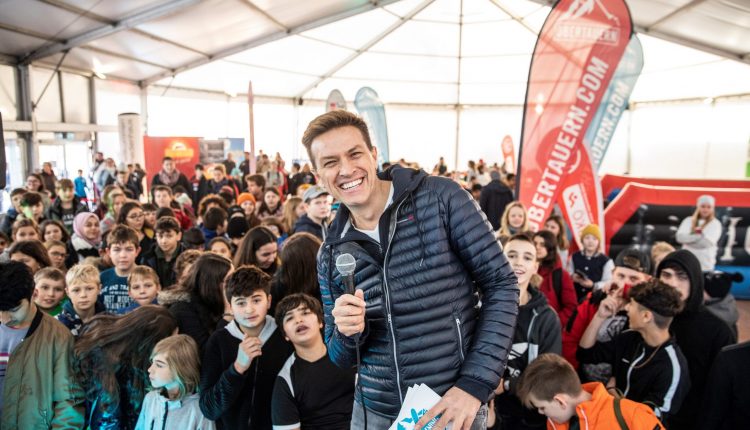 Ein Mann mit einem Mikrofon lächelt bei einer überfüllten Veranstaltung in einer Halle mit vielen Kindern und bunten Bannern.