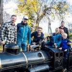 Acht Personen posieren und lächeln um eine schwarze Miniatur-Dampfeisenbahn in einem Park mit Herbstbäumen.
