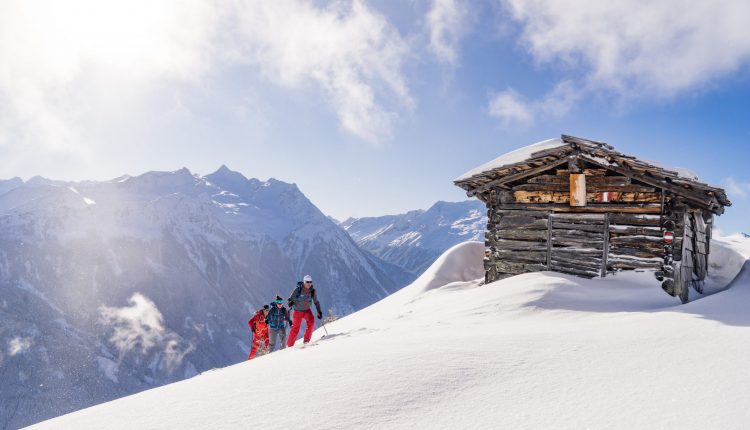Zwei Personen wandern im Tiefschnee bergauf in der Nähe einer Holzhütte, mit verschneiten Bergen im Hintergrund.