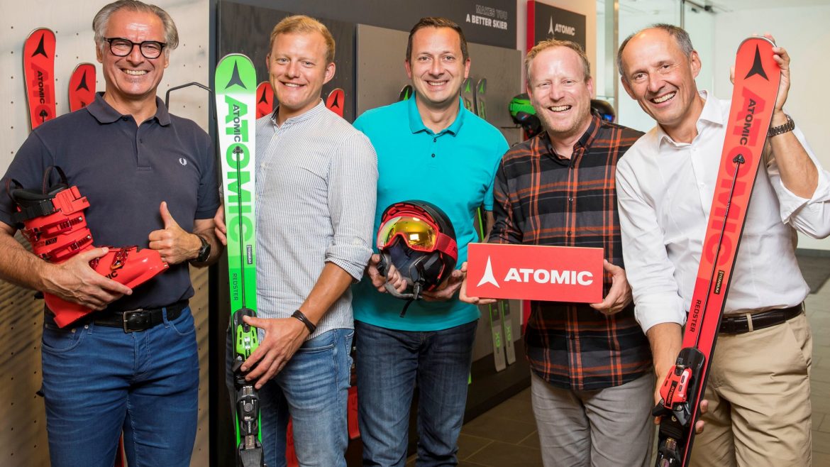 Fünf lächelnde Männer halten Atomic-Ski und -Ausrüstung in der Hand und stehen gemeinsam in einem Raum vor einer Ausstellungswand.