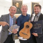 Drei lächelnde Männer posieren mit einer Gitarre, CDs und einer Schallplatte vor gerahmten Porträts.