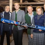 Sechs lächelnde und posierende Menschen, die einen blauen Ski unter einem Holzunterstand halten.