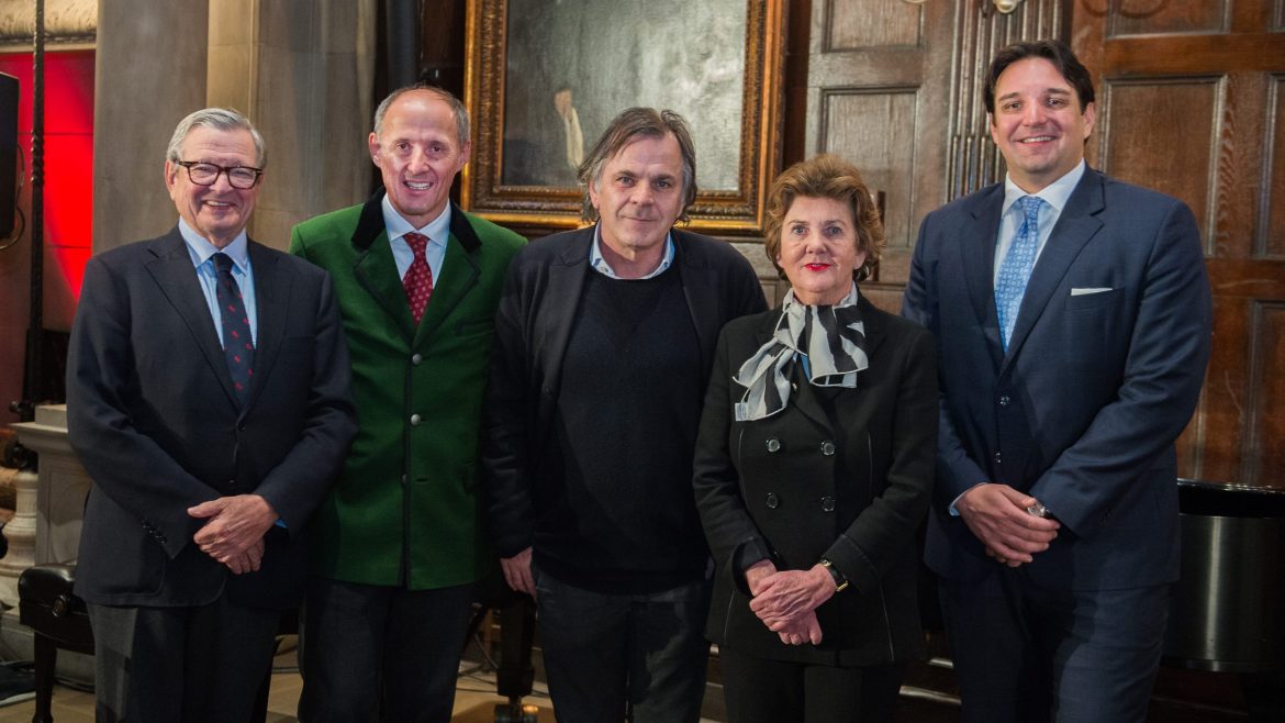 Fünf formell gekleidete Personen stehen in einem Innenraum zusammen und lächeln für ein Gruppenfoto vor einer holzgetäfelten Wand.