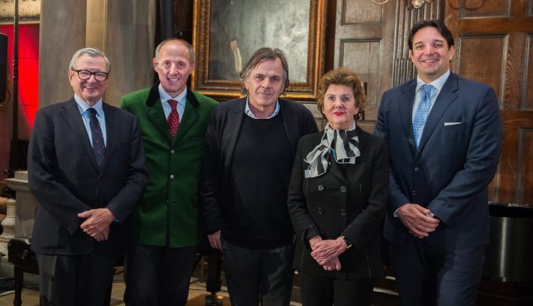 Fünf formell gekleidete Personen stehen in einem Innenraum zusammen und lächeln für ein Gruppenfoto vor einer holzgetäfelten Wand.