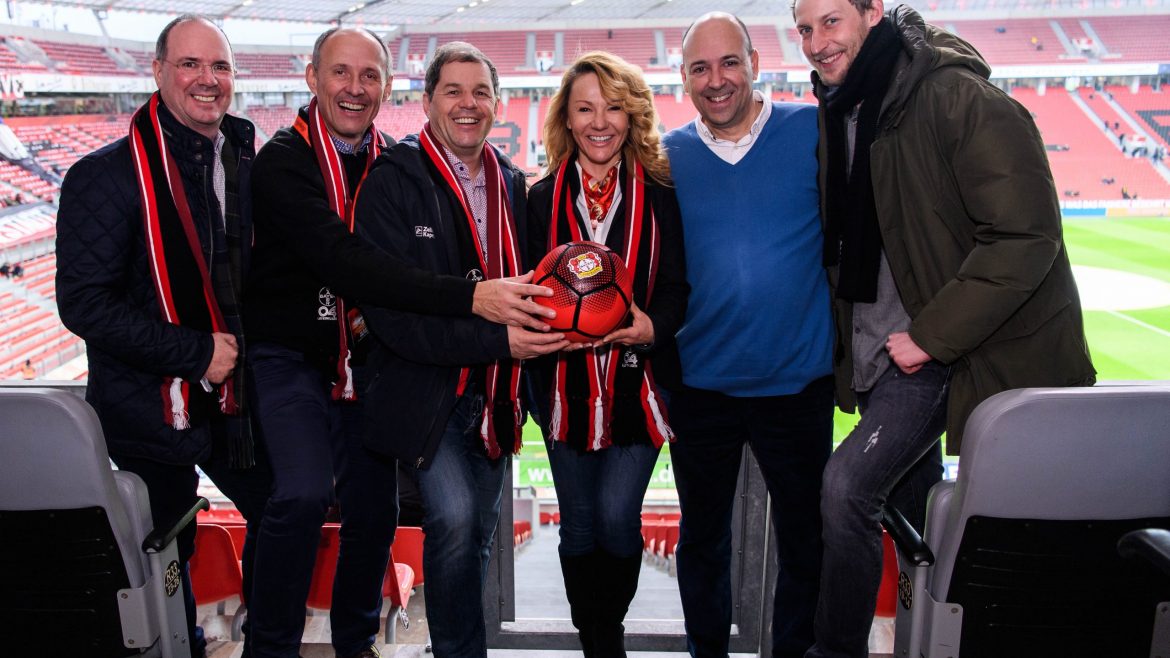 Sechs lächelnde Menschen mit Schals posieren zusammen und halten einen Fußball in einem Stadion mit Blick auf das Spielfeld.