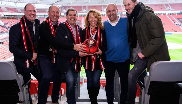 Sechs lächelnde Menschen mit Schals posieren zusammen und halten einen Fußball in einem Stadion mit Blick auf das Spielfeld.