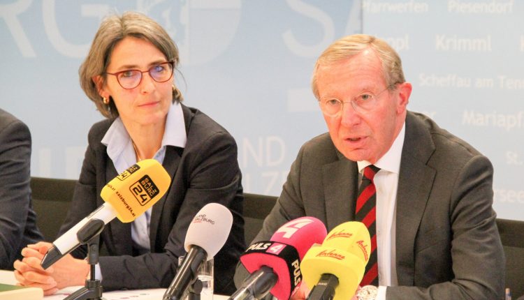 Eine Frau und ein Mann in Geschäftskleidung sprechen auf einer Pressekonferenz mit mehreren Mikrofonen vor ihnen.