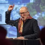 Ein älterer Mann spricht leidenschaftlich in ein Mikrofon und gestikuliert mit einer Hand, die er über ein offenes Buch hält.