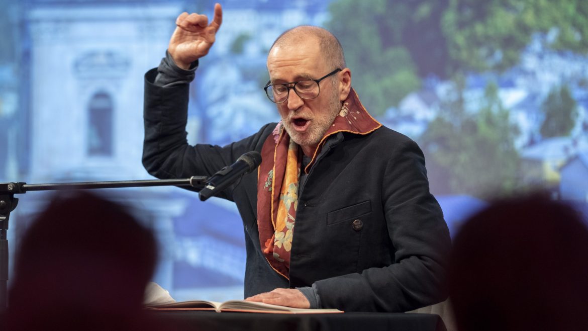 Ein Mann mit Brille liest leidenschaftlich in ein Mikrofon und hebt eine Hand zur Betonung.