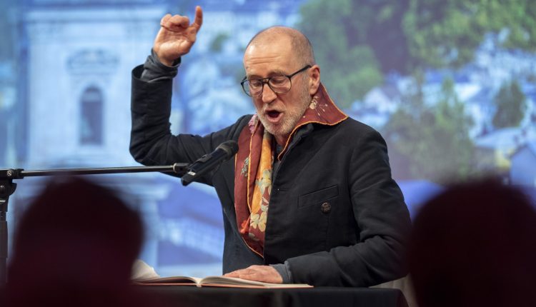 Ein Mann mit Brille liest leidenschaftlich in ein Mikrofon und hebt eine Hand zur Betonung.