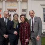 Vier Erwachsene in formeller Kleidung posieren lächelnd vor einem Gebäude mit der Aufschrift "Theater im Zimmer".