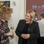 Zwei Frauen unterhalten sich lächelnd bei einer Indoor-Veranstaltung, eine hält ein Getränk in der Hand, mit Menschen und Kunst im Hintergrund.