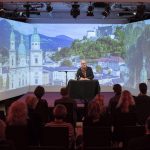 Ein Redner wendet sich an ein Publikum in einem Raum, in dem hinter ihm szenische Stadtbilder projiziert werden.