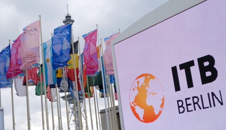 Bunte internationale Flaggen wehen neben einem großen ITB Berlin-Schild und einem Kommunikationsturm unter einem bewölkten Himmel.