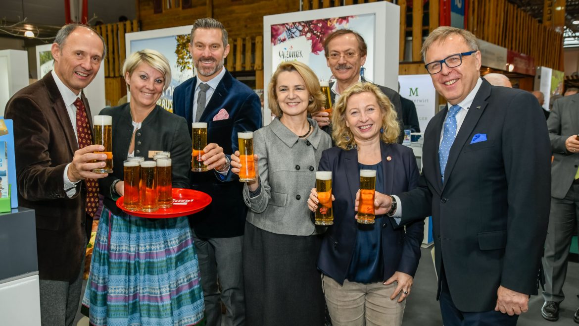 Sieben Personen in Geschäftskleidung lächeln und halten Biergläser bei einer Indoor-Veranstaltung.