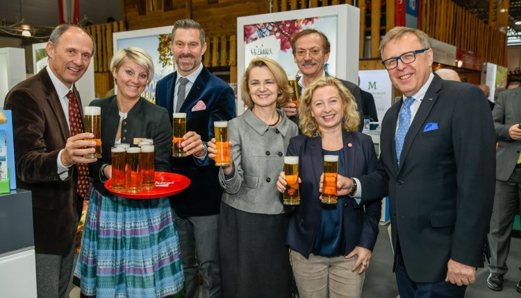 Sieben Personen in Geschäftskleidung lächeln und halten Biergläser bei einer Indoor-Veranstaltung.