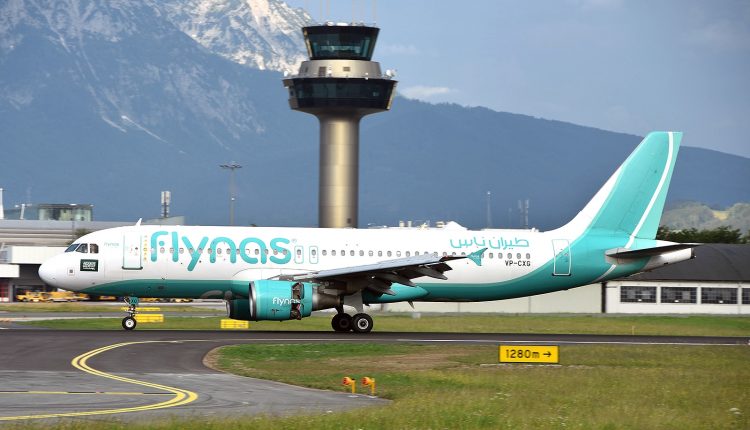 Ein Flynas-Flugzeug rollt auf einer Landebahn mit einem Kontrollturm und Bergen im Hintergrund.