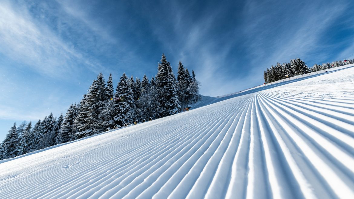 Präparierte Skipiste mit parallelen Linien, verschneiten Bäumen und einem blauen Himmel mit Wolkenfetzen.