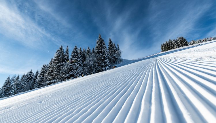 Präparierte Skipiste mit parallelen Linien, verschneiten Bäumen und einem blauen Himmel mit Wolkenfetzen.