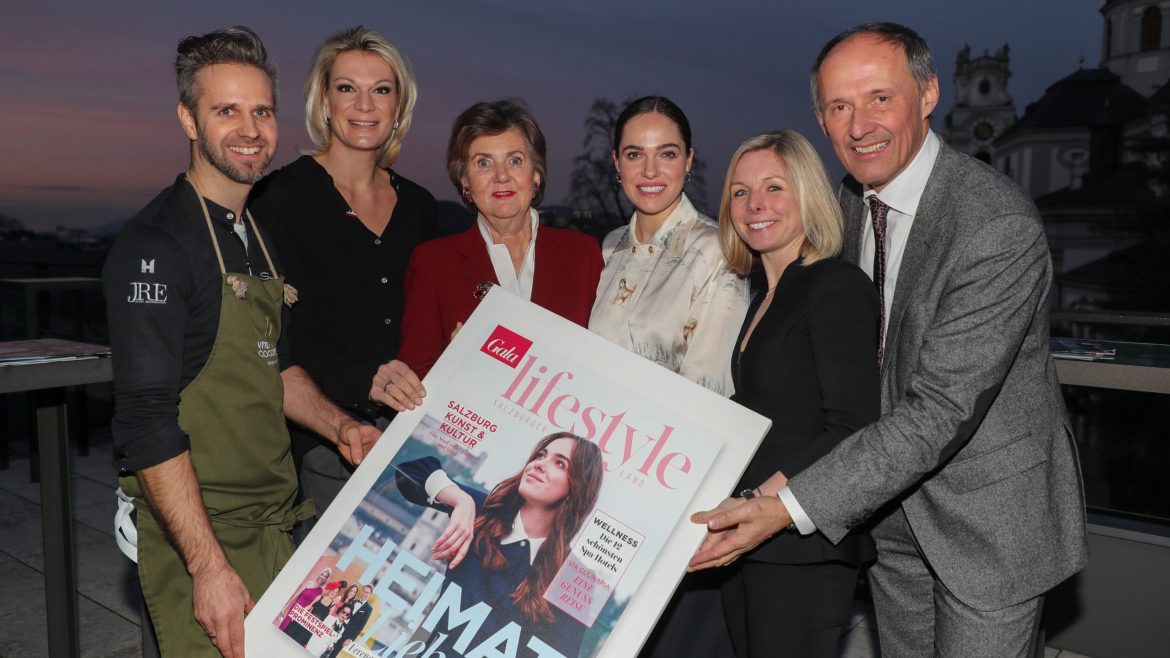 Sechs Personen posieren bei Sonnenuntergang im Freien und halten ein großes Lifestyle-Magazin, auf dem eine Frau abgebildet ist.