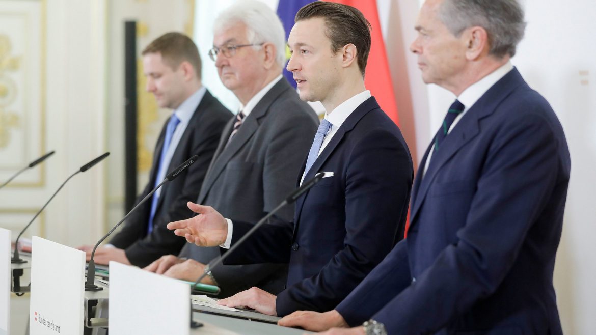 Vier Männer in Anzügen stehen auf einem Podium und sprechen auf einer formellen Pressekonferenz mit Fahnen im Hintergrund.