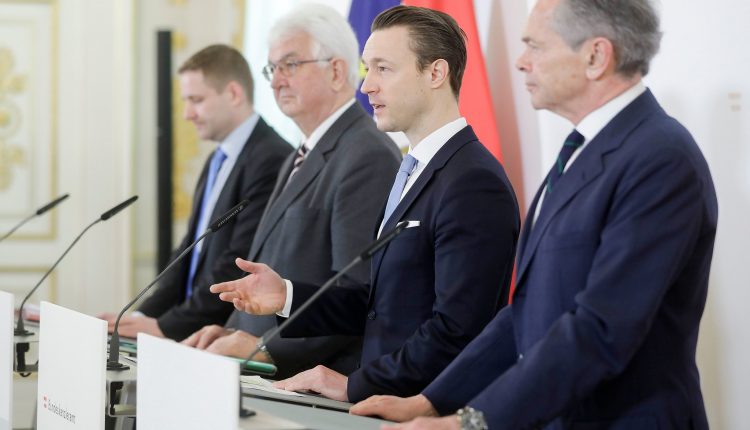Vier Männer in Anzügen stehen auf einem Podium und sprechen auf einer formellen Pressekonferenz mit Fahnen im Hintergrund.