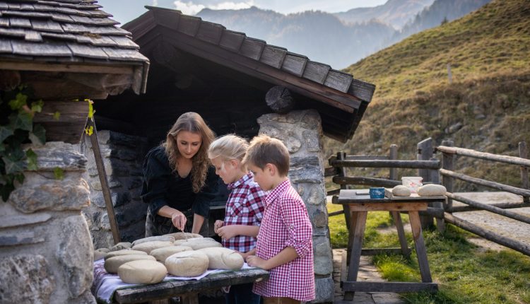 Eine Frau und zwei Kinder kneten draußen vor einem Steinschuppen in einer Berglandschaft Teig.