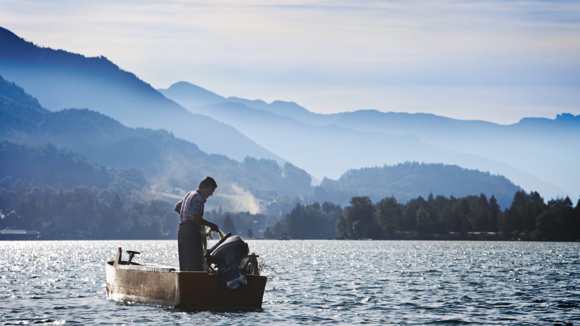 Eine Person steht in einem kleinen Boot auf einem See, umgeben von nebligen Bergen unter blauem Himmel.