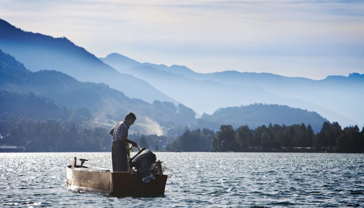 Eine Person steht in einem kleinen Boot auf einem See, umgeben von nebligen Bergen unter blauem Himmel.