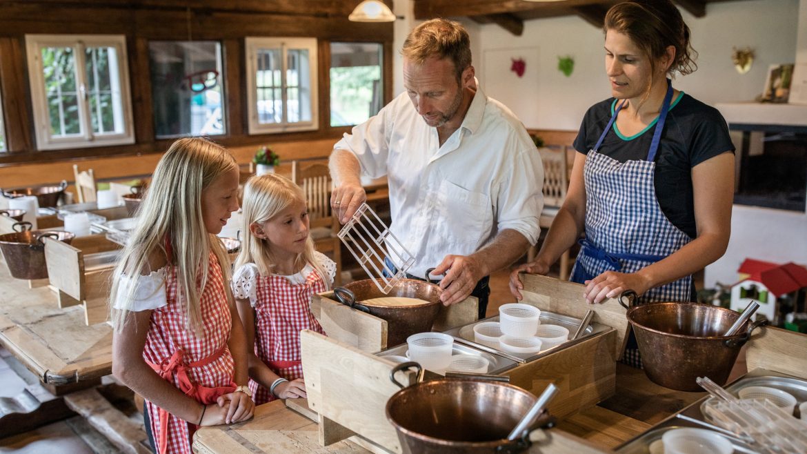Ein Mann und eine Frau zeigen zwei Mädchen, wie man in einer rustikalen Küche kocht, alle tragen Schürzen.