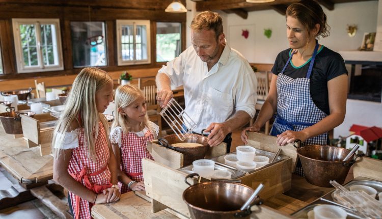 Ein Mann und eine Frau zeigen zwei Mädchen, wie man in einer rustikalen Küche kocht, alle tragen Schürzen.