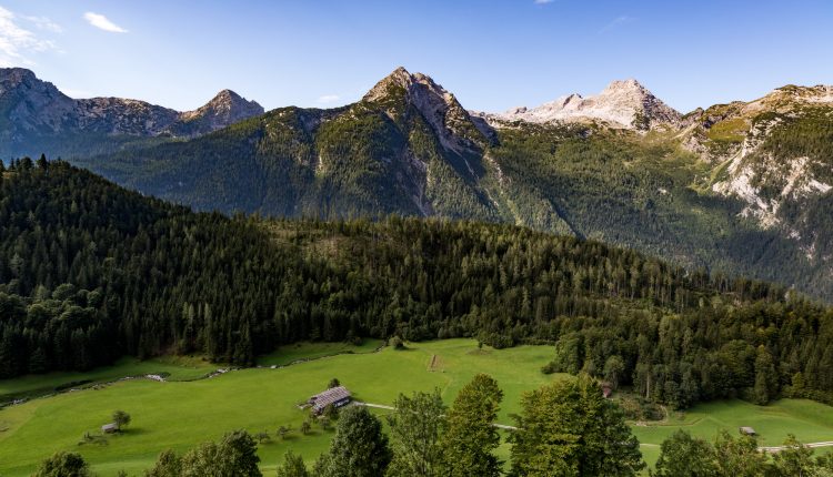 Ein grünes Tal mit verstreuten Hütten, dichten Kiefernwäldern und hohen Bergen unter einem klaren blauen Himmel.