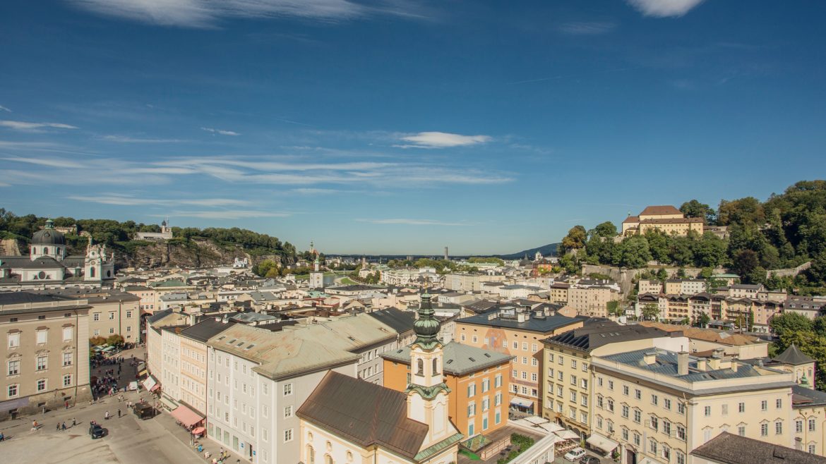 Luftaufnahme von Salzburg, Österreich, mit farbenfrohen historischen Gebäuden und Hügeln unter einem klaren blauen Himmel.