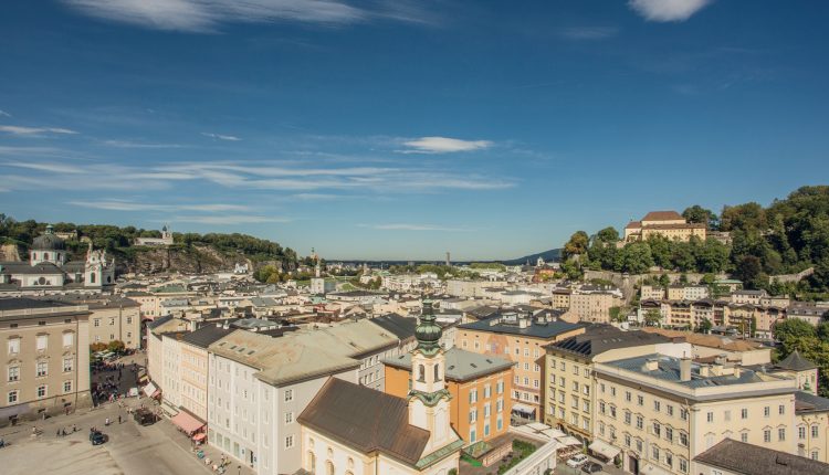 Luftaufnahme von Salzburg, Österreich, mit farbenfrohen historischen Gebäuden und Hügeln unter einem klaren blauen Himmel.