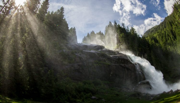 Ein Wasserfall ergießt sich in Kaskaden durch einen Wald, während die Sonnenstrahlen unter einem blauen Himmel durch hohe Kiefern scheinen.