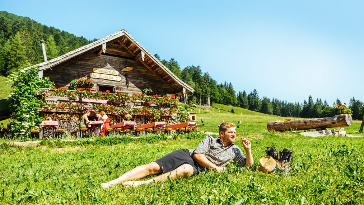 Ein Mann entspannt sich im Gras vor einer blumengeschmückten Holzhütte in einer sonnigen, ländlichen Berglandschaft.
