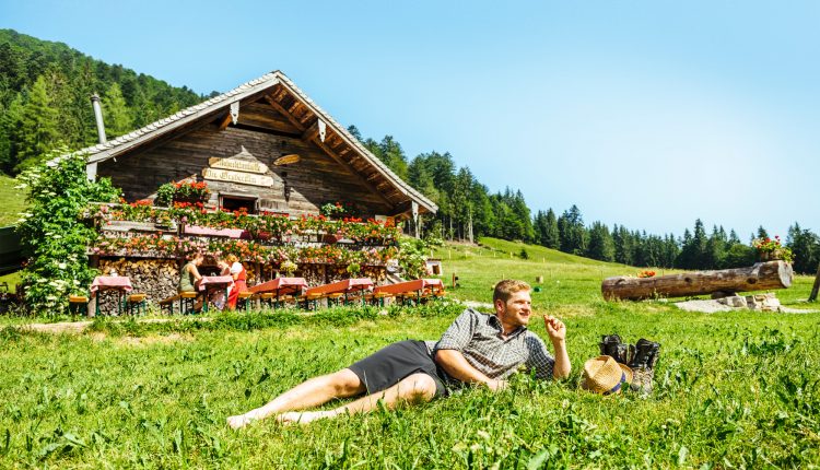 Ein Mann entspannt sich im Gras vor einer blumengeschmückten Holzhütte in einer sonnigen, ländlichen Berglandschaft.