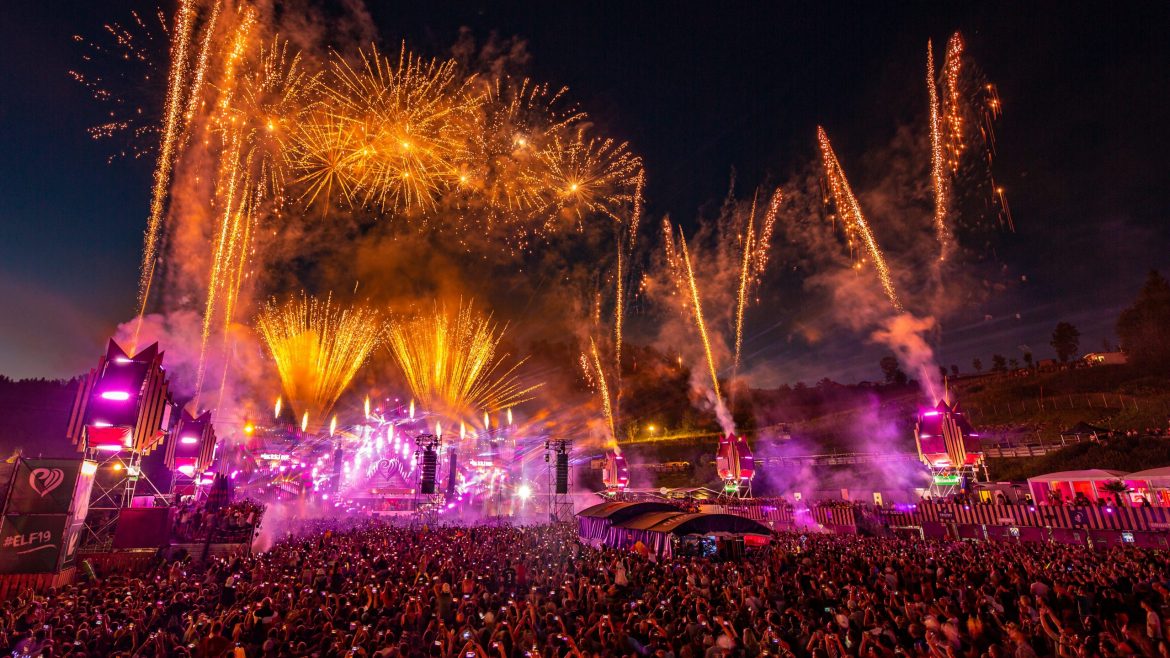 Eine große Menschenmenge bei einem nächtlichen Musikfestival mit Feuerwerk und bunten Bühnenlichtern am Himmel.