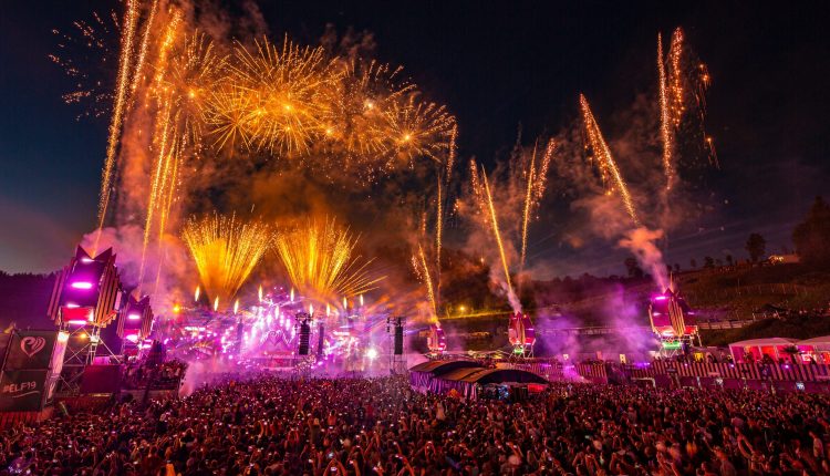 Eine große Menschenmenge bei einem nächtlichen Musikfestival mit Feuerwerk und bunten Bühnenlichtern am Himmel.