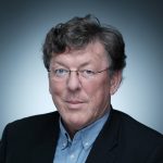 Ein Mann mit Brille und lockigem Haar, der ein blaues Hemd und einen dunklen Blazer trägt, posiert vor einem Hintergrund mit Farbverlauf.