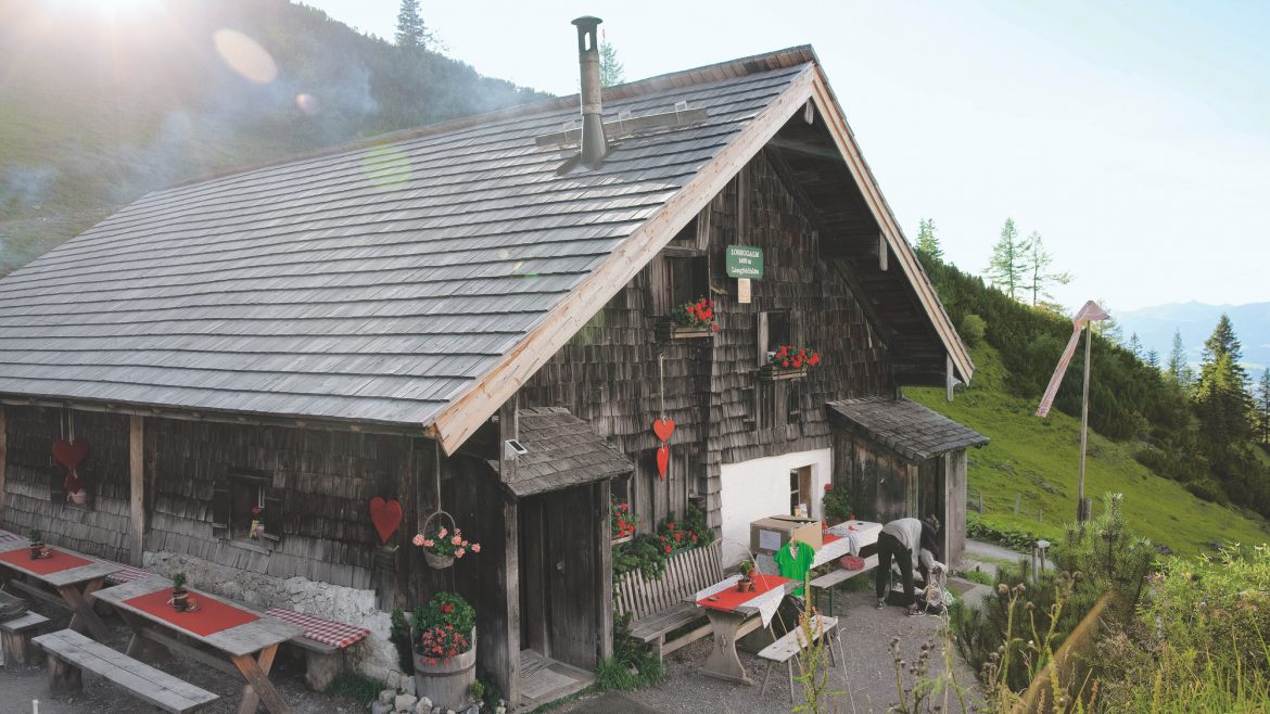 Eine rustikale hölzerne Berghütte mit Sitzgelegenheiten im Freien, roten Akzenten und einer malerischen Grünanlage im Hintergrund.