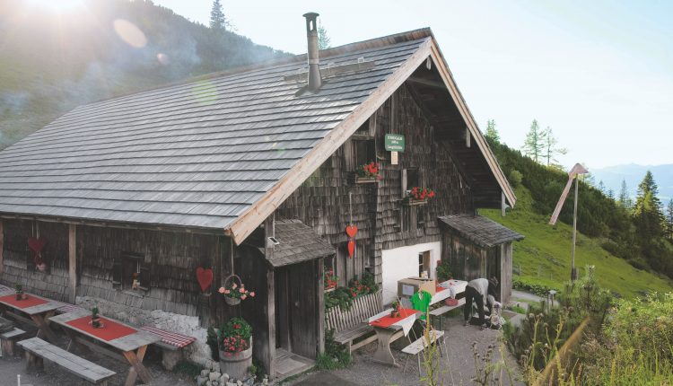 Eine rustikale hölzerne Berghütte mit Sitzgelegenheiten im Freien, roten Akzenten und einer malerischen Grünanlage im Hintergrund.